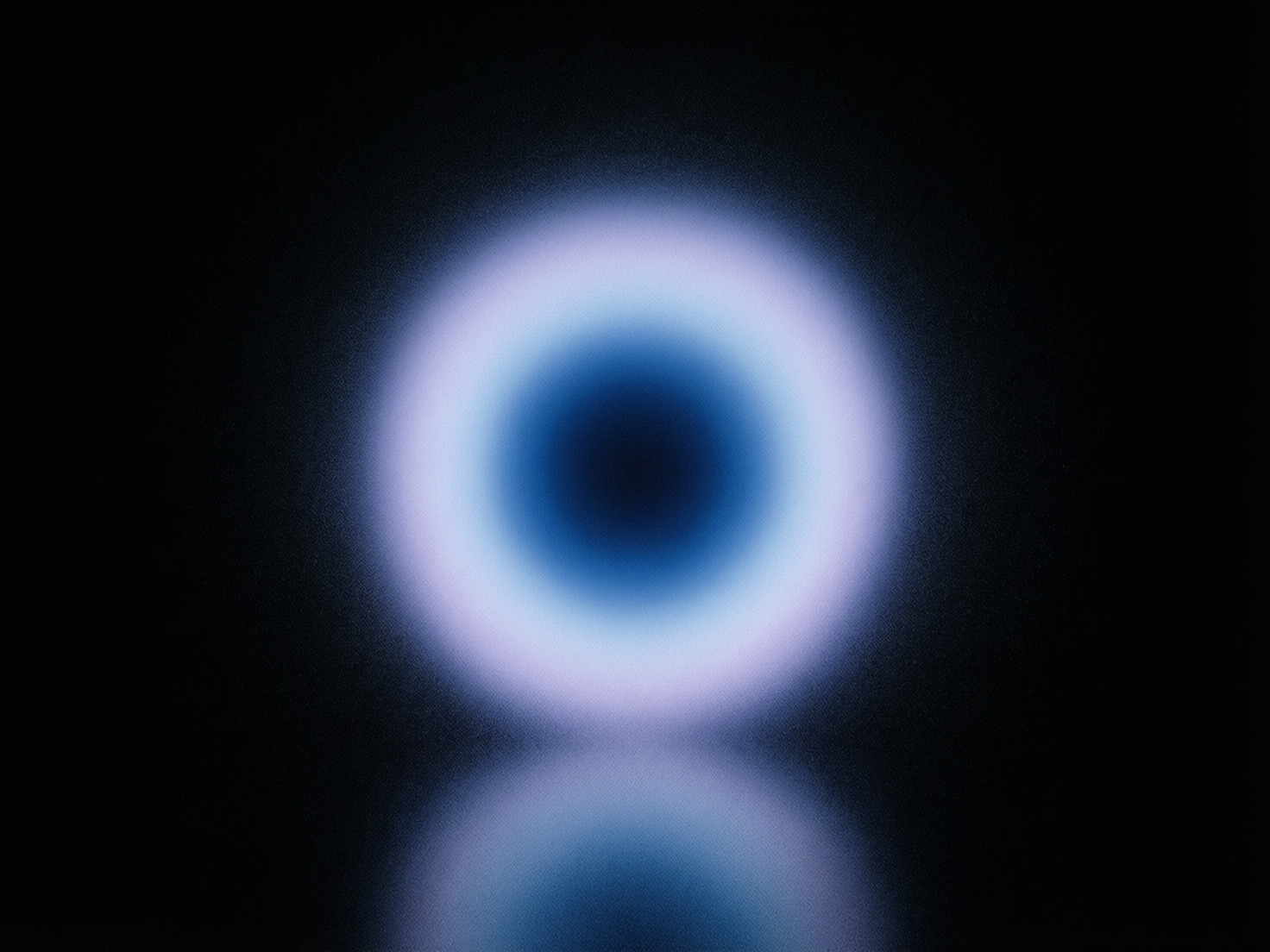 图中展示了一个极简主义风格的抽象发光圆环，悬浮在深邃的黑色背景中心。这个圆环呈现出柔和的渐变效果，中心是一个深黑色的圆洞，向外依次扩散为深蓝色、亮蓝色，最外层则是带有淡淡紫色调的白色光晕。整个光圈具有明显的颗粒感和磨砂质感，边缘模糊，仿佛一种虚幻的能量场。在圆环下方，有一个模糊的倒影，暗示着下方存在一个光滑的反射平面。

从 AI 生成的角度来看，画面构图非常对称，光影过渡自然，但在细节上存在一些特征：光圈的颗粒分布略显机械化，缺乏自然光学的随机性。此外，下方的倒影虽然在位置上与主体对应，但其形状的拉伸感与上方的正圆形并不完全符合物理透视规律，显得有些生硬。整体而言，这是一张充满未来感和冥想氛围的视觉艺术图。