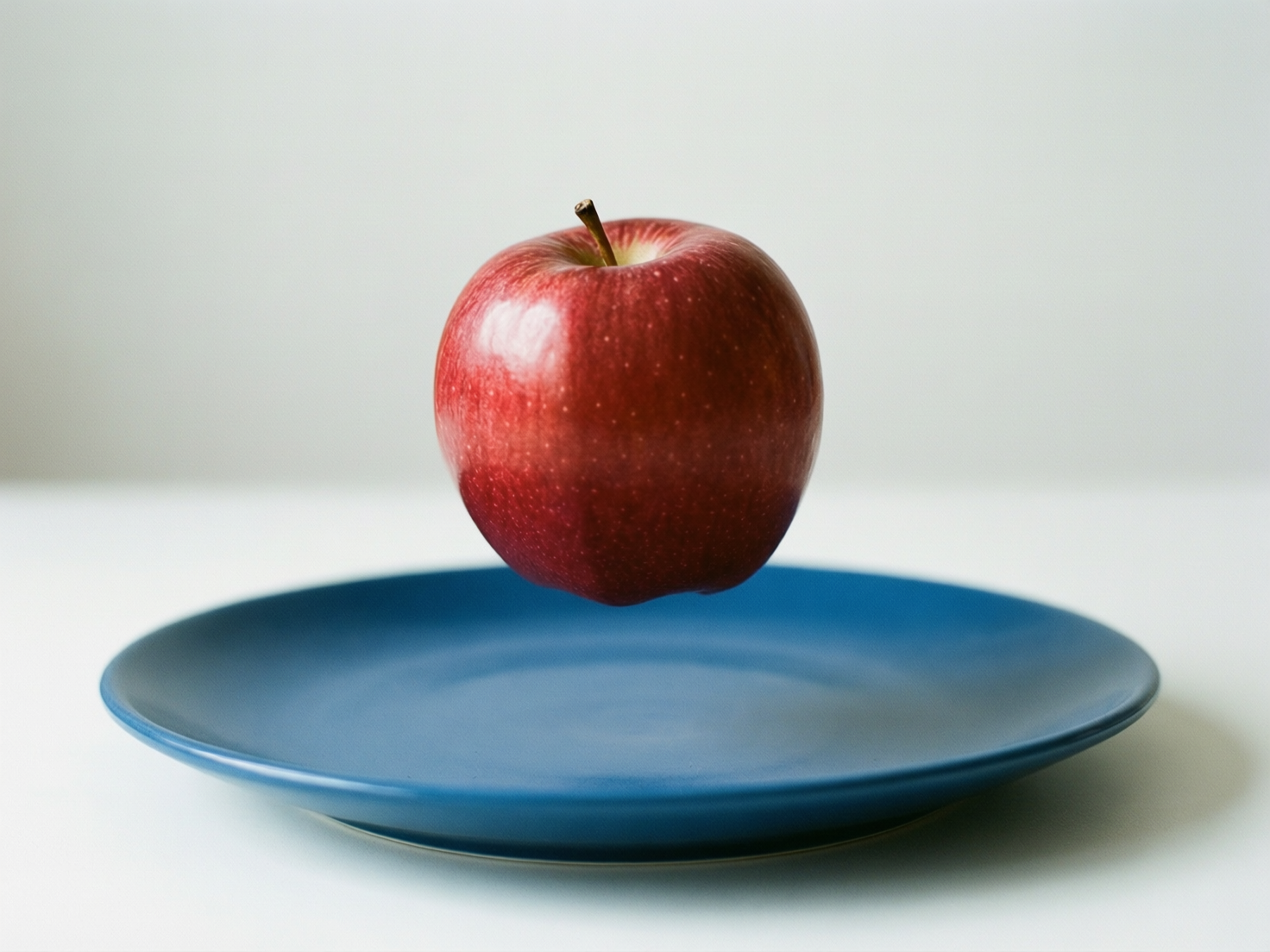 图中展示了一个极简主义风格的静物场景。画面中心是一个硕大、饱满的红苹果，它正神奇地悬浮在一个深蓝色圆盘的上方。苹果表皮呈现出深浅不一的红色，带有细小的浅色斑点，左侧有一抹明显的反光，显得质感清脆。下方的蓝色盘子质地细腻，呈现出哑光质感，平稳地放置在纯白色的桌面上。背景是干净的浅灰色墙面，整体色调明快且对比鲜明。

在 AI 生成的细节方面，存在明显的物理逻辑缺陷：苹果在没有任何支撑的情况下悬浮，且盘面上完全没有出现苹果应有的投影，这种光影关系的缺失使得苹果看起来像是后期粘贴上去的。此外，苹果顶部的果柄与果肉连接处略显模糊，盘子的边缘轮廓在右侧也显得有些不够自然。这种超现实的构图虽然视觉冲击力强，但违背了现实世界的重力与光学规律。