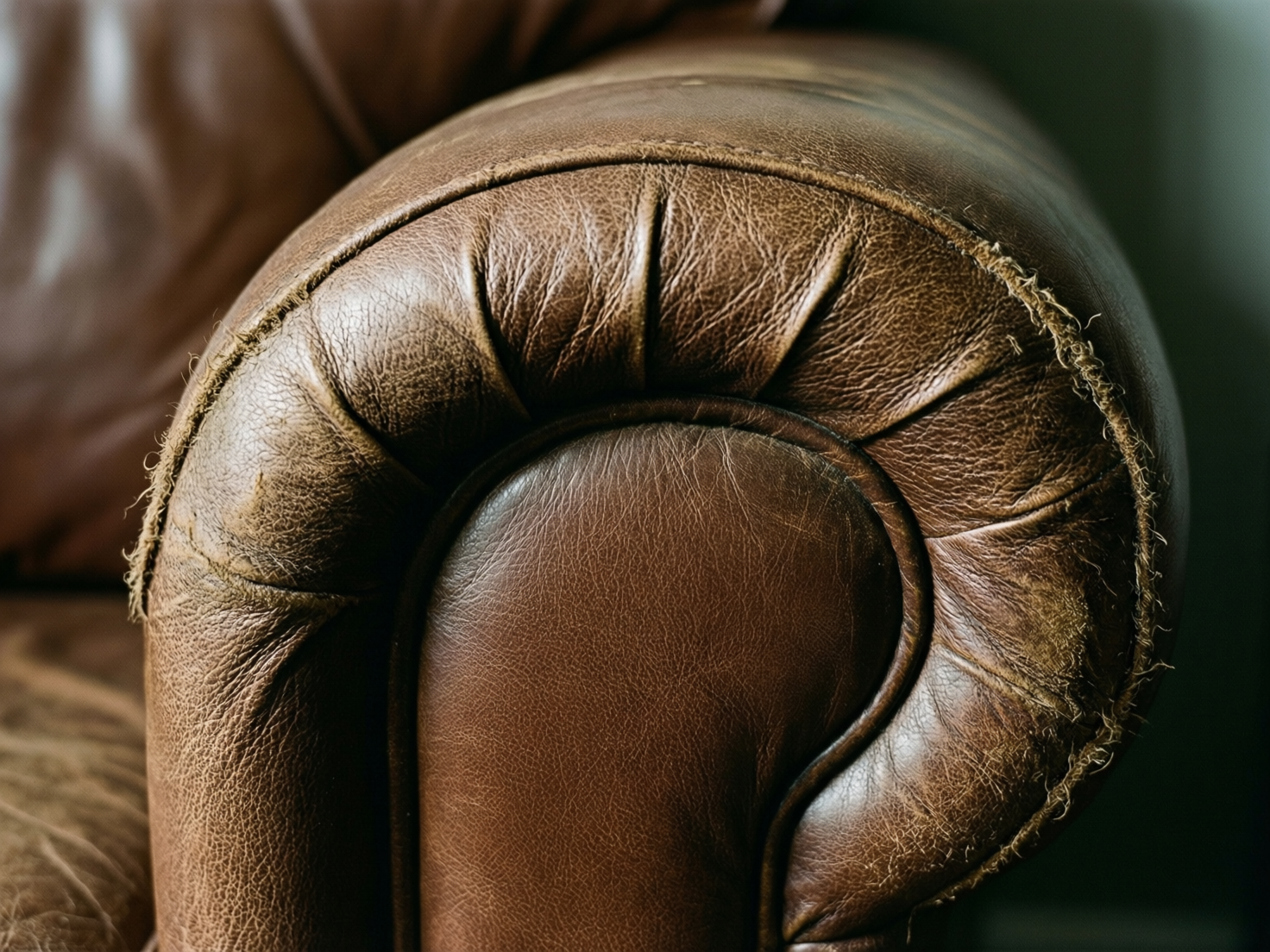图中展示了一张棕色复古皮质沙发的扶手特写。画面以暖色调为主，皮革呈现出深浅不一的焦糖色，表面布满了细腻的纹理和自然的褶皱，展现出一种陈旧而厚重的质感。扶手的造型圆润，边缘带有典型的切斯特菲尔德风格折叠缝线。光线从右上方照射，在皮革表面形成了柔和的高光与深邃的阴影。

在 AI 生成的细节方面，画面存在明显的逻辑缺陷：扶手边缘的缝线处出现了异常的磨损和纤维拉丝，这些毛边看起来更像是粗糙的麻绳或生物组织，而非真实的皮革纤维，且分布极不自然。此外，扶手内侧的圆弧缝合线在底部交汇处显得结构模糊，纹路走向略显混乱，缺乏真实家具制造中的严谨工艺感。背景部分被大面积虚化，只能隐约分辨出沙发的靠背阴影。