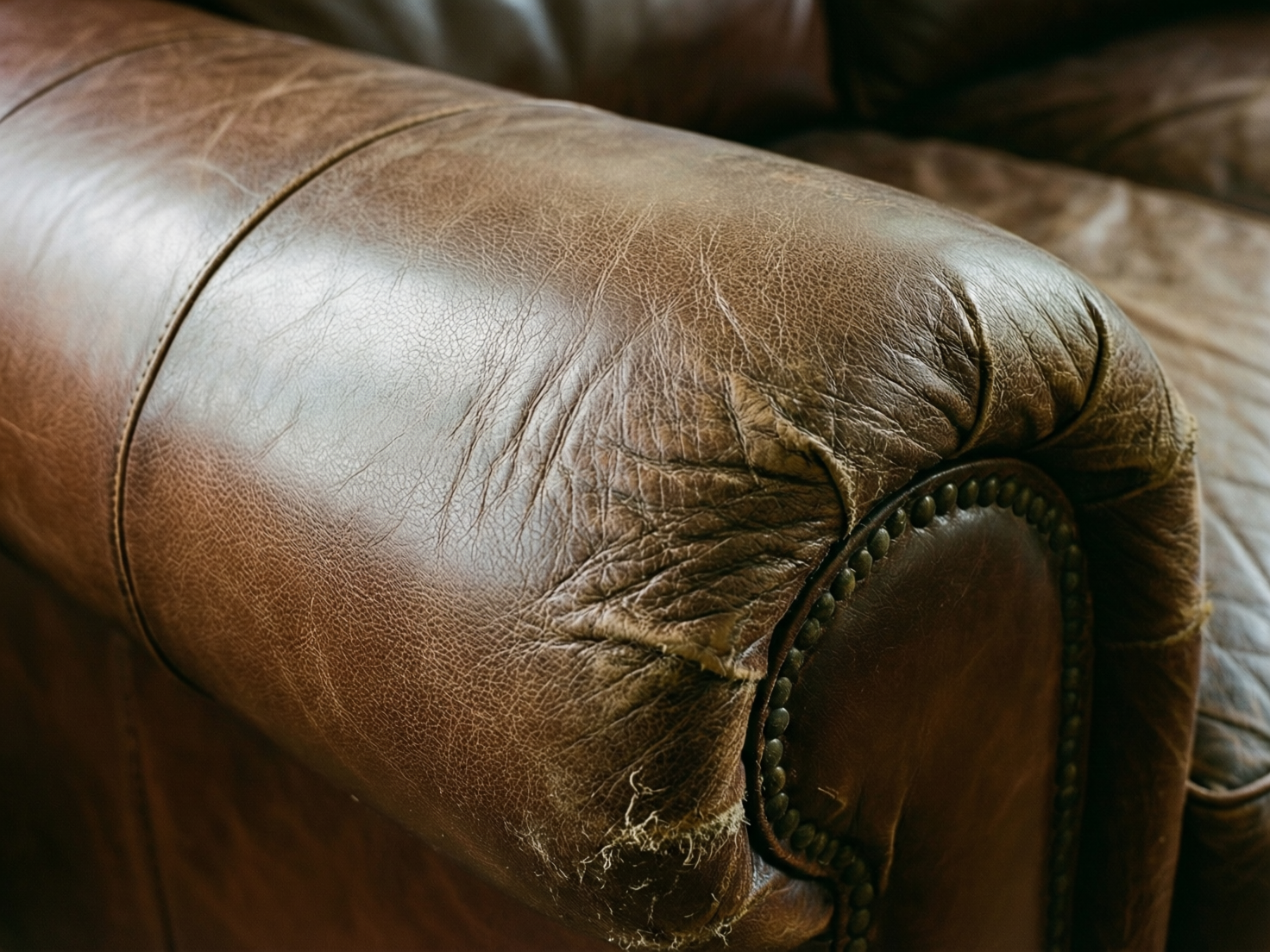 图中展示了一张棕色复古皮质沙发的扶手细节特写。画面色调沉稳，光线从左上方照射，在皮革表面形成了柔和的高光。扶手呈现圆柱状，皮革纹理清晰可见，布满了细密的褶皱和自然的磨损痕迹。扶手的侧面边缘装饰有一圈古铜色的金属铆钉，增强了古典家具的质感。然而，画面中存在明显的 AI 生成缺陷：在扶手正前方的皮革接缝处，出现了不自然的撕裂和模糊感，一些纤维状的线条杂乱地悬浮在半空，未能与皮革材质完美融合。此外，右侧背景中的沙发靠背线条略显扭曲，透视关系不够严谨。整体构图集中在扶手的转角处，通过细腻的质感表现出一种陈旧而温暖的居家氛围。