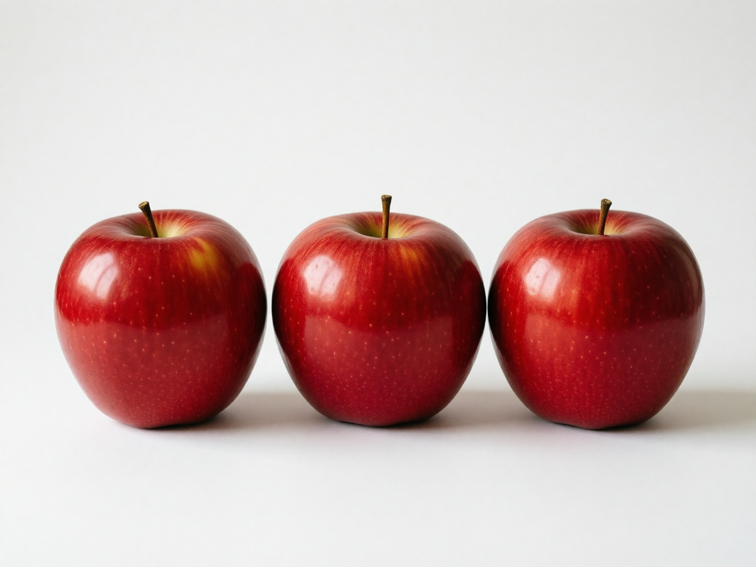 图中展示了三个鲜红色的苹果，它们整齐地横向排列在纯白色的背景中央。这些苹果色泽饱满，果皮上带有细小的浅色斑点，表面反射着柔和的光泽，展现出一种写实的质感。每个苹果的顶部都有一根短小的深褐色果柄。构图非常简洁、对称，光线从左上方照射，在苹果右侧的白色平面上留下了淡淡的阴影。

作为一张 AI 生成的图片，画面在细节处理上存在一些不自然之处。首先，三个苹果的形状、大小以及表面的高光位置几乎完全一致，这种高度的重复性在自然界中非常罕见，显得有些机械。其次，苹果底部的阴影处理过于模糊且生硬，缺乏真实物体与平面接触时应有的细腻过渡。此外，最左侧苹果的果柄连接处显得有些模糊，与果肉的融合不够自然，呈现出一种拼贴感。